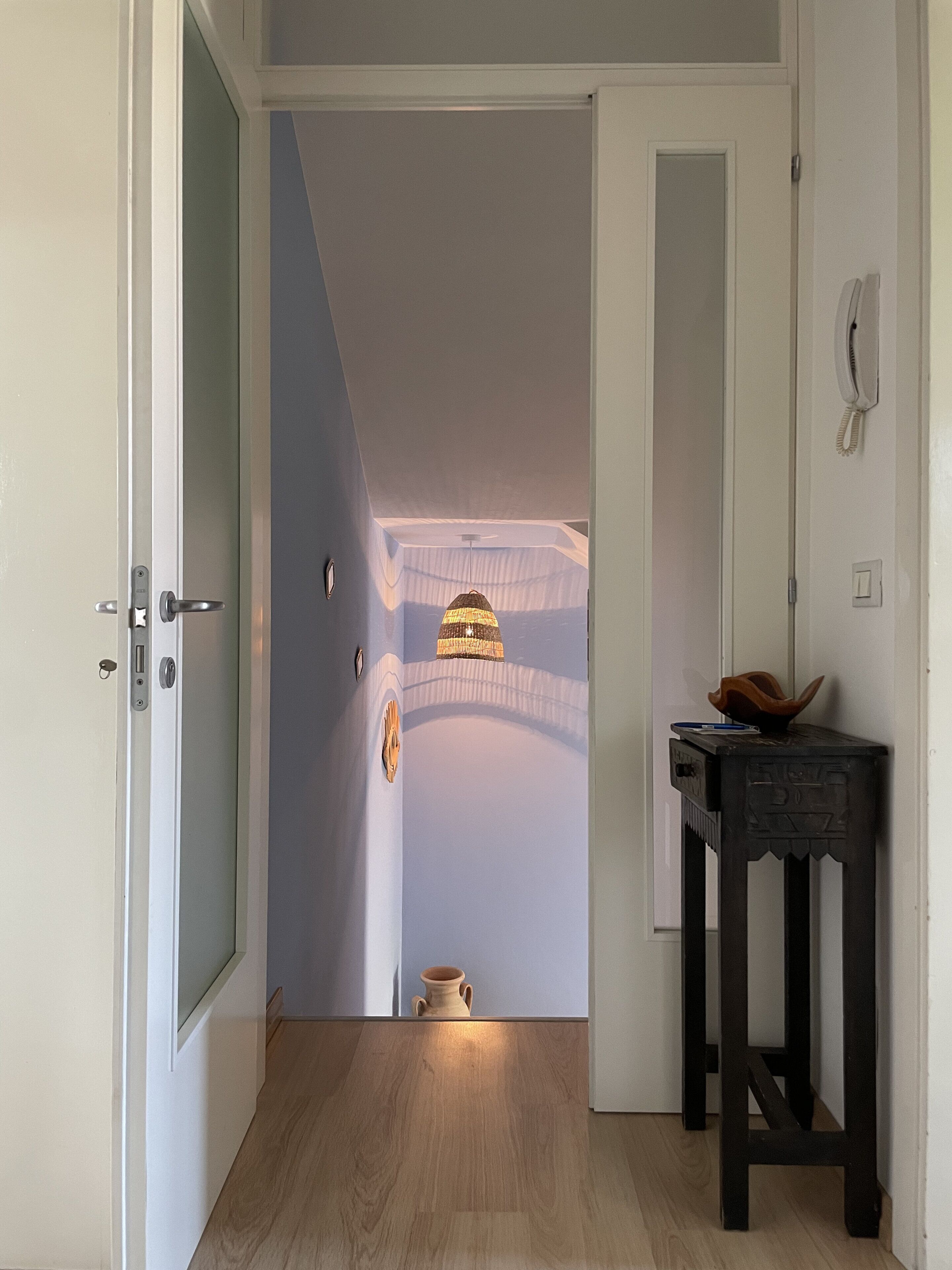 Un corridoio con pavimento in legno, una porta sul lato sinistro, un piccolo tavolo con un vaso a destra e un lampadario al soffitto.