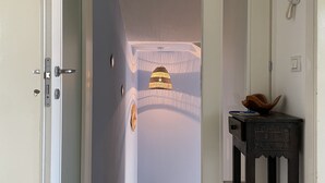 Um corredor com piso de madeira, porta à esquerda, uma pequena mesa com um vaso à direita e uma luminária no teto.
