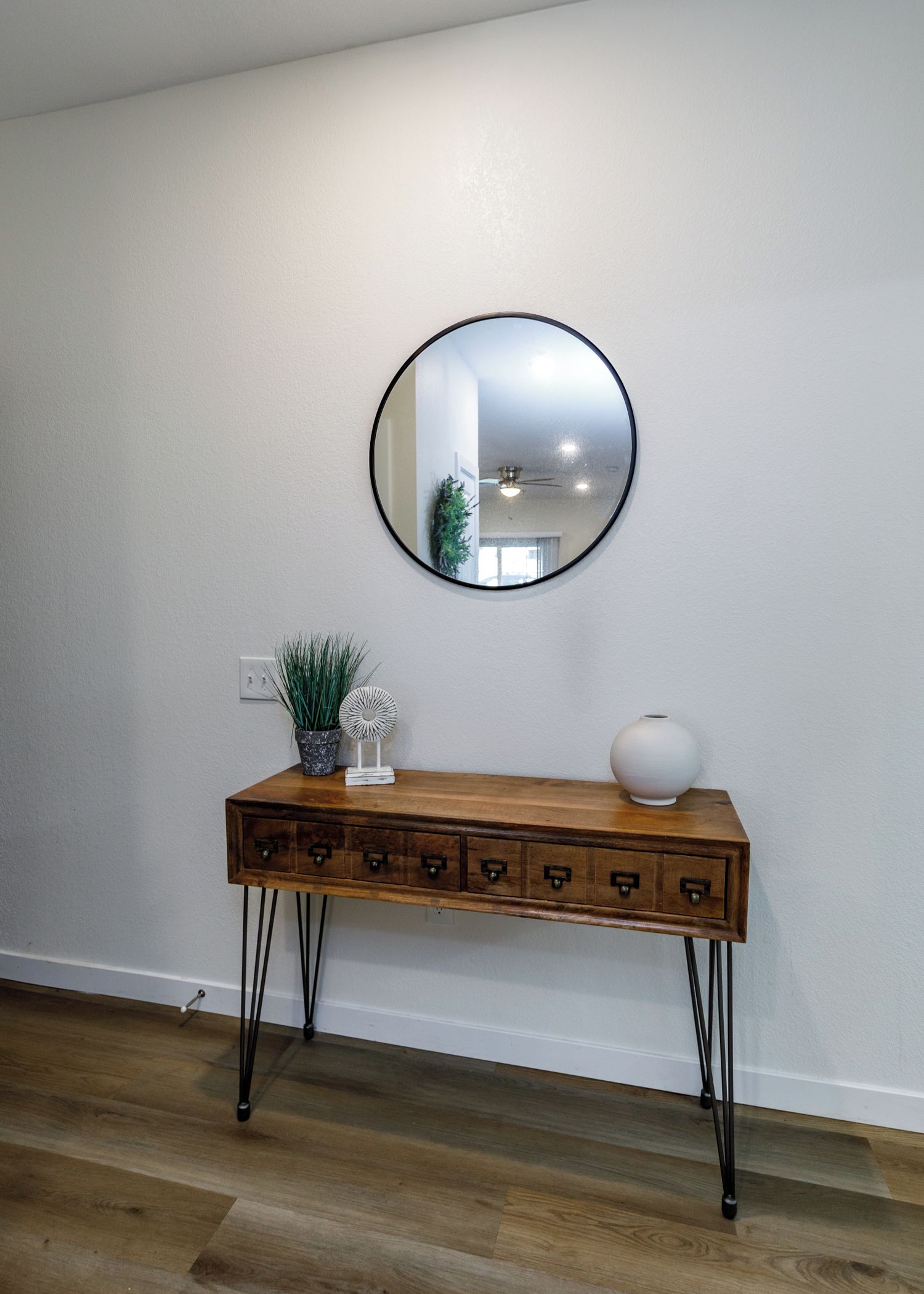 Una consola de madera con cajones, un espejo redondo, una planta en maceta y un florero frente a una pared blanca.