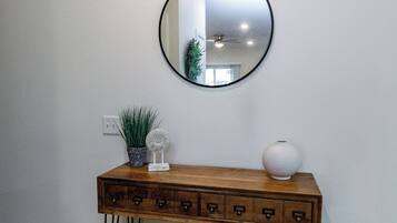 Una consola de madera con cajones, un espejo redondo, una planta en maceta y un florero frente a una pared blanca.