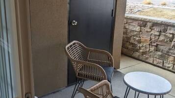 A balcony with wicker chairs and a small table, a dark door, and a view of a residential area.