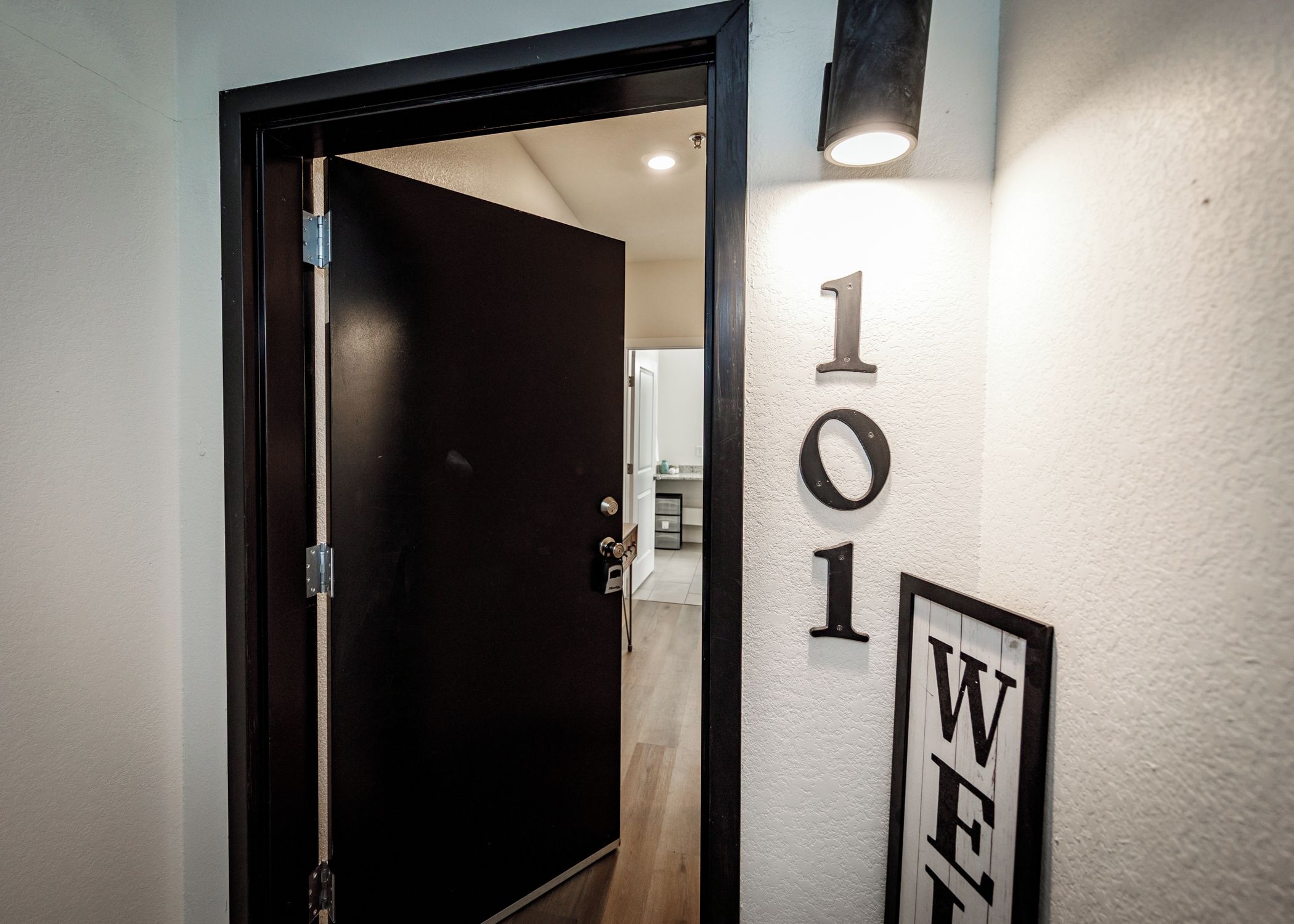 Una puerta de madera oscura con un cierre de metal, una pared blanca con el número '101' y un letrero con la letra 'W', además de una lámpara empotrada.