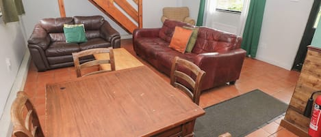 A living room with a wooden table and chairs, two leather sofas, a wooden staircase, and a window with curtains.