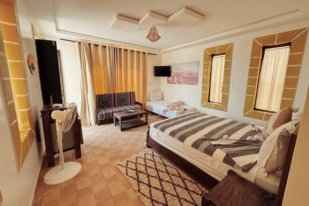 Quarto com cama, sofá, televisão, ventilador e janela com cortinas.