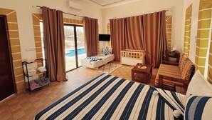 A bedroom with a bed, a wooden crib, a sofa, a TV, and a view of a swimming pool. - Villa Ryad Ibou (Aguerd)