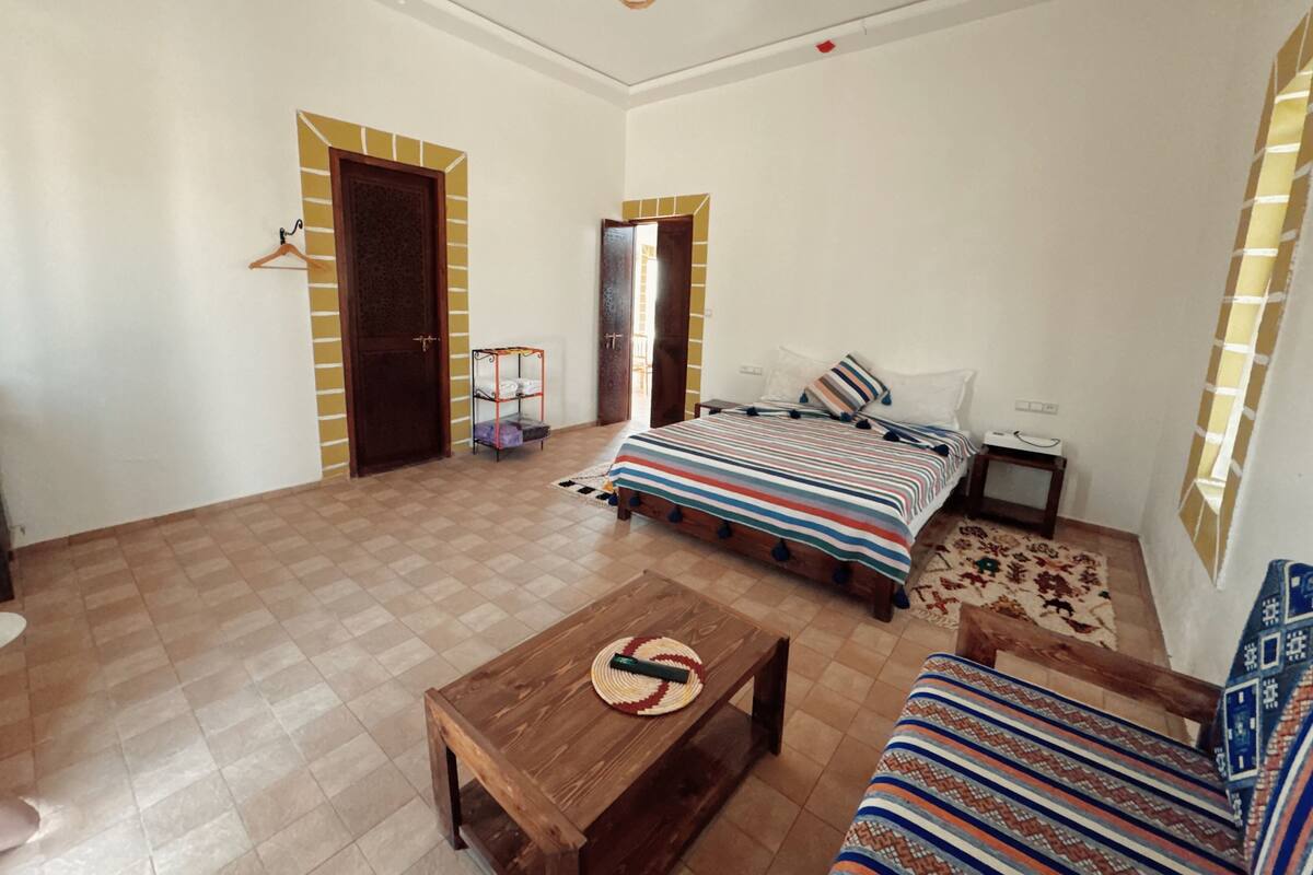 Quarto com cama, mesa de madeira, sofá, luminária pendente e uma porta.