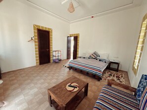 A room with a bed, a wooden table, a sofa, a hanging light fixture, and a door.