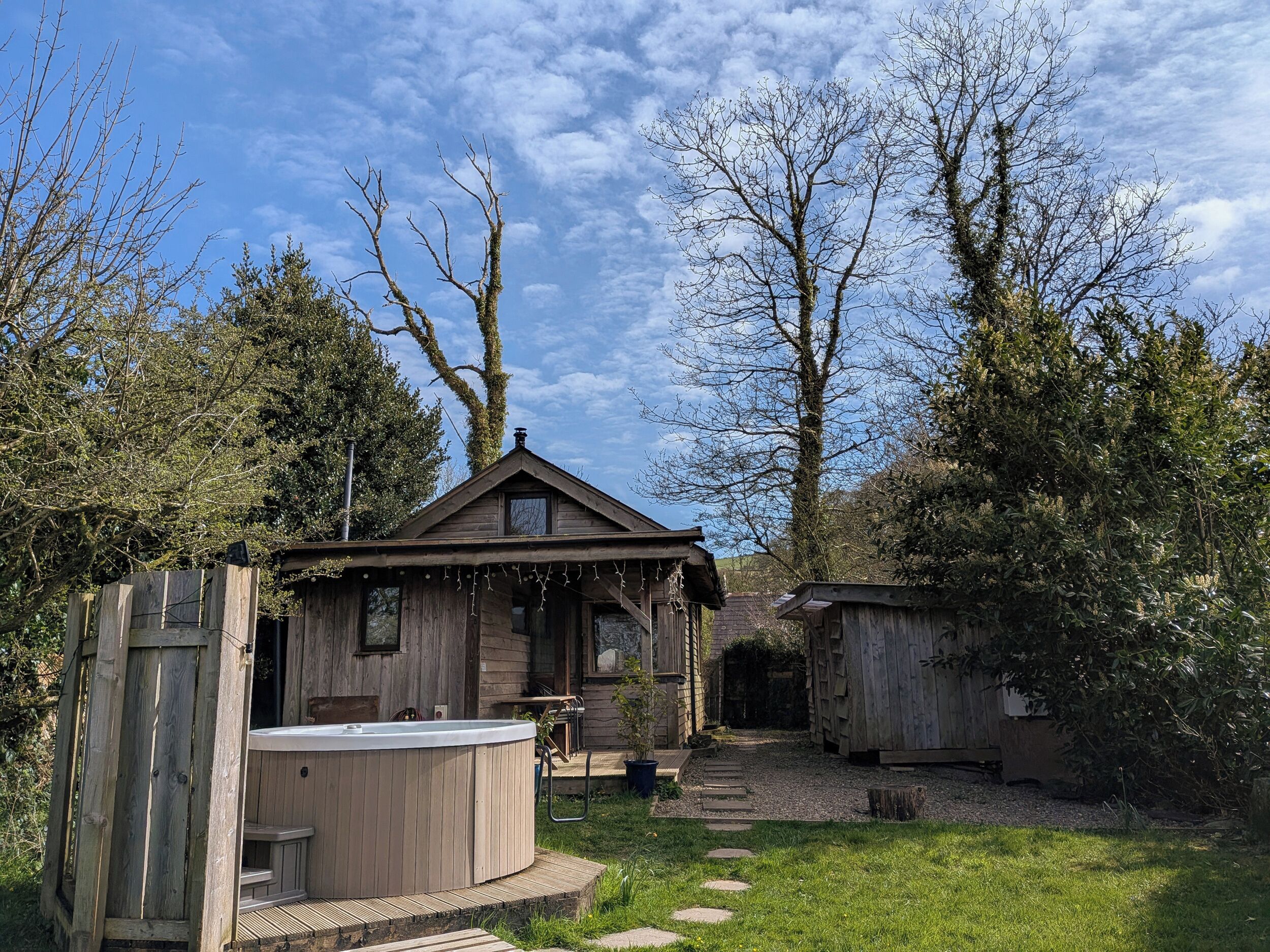 A wooden cabin with a hot tub, surrounded by trees and a gravel path.