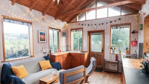 A cozy cabin interior with a wooden ceiling, a sofa with cushions, a dining table, a kitchen area, and large windows. - Cosy Eco Cabin (Newport)