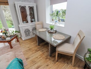 Un coin repas comprenant une table à plateau de verre, des chaises en osier, un buffet blanc aux portes vitrées et une fenêtre donnant sur un paysage verdoyant.