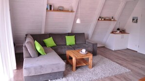 A loft-style living room with a gray sectional sofa, green cushions, a wooden coffee table, and white walls with exposed beams. - Holiday Home 'Meinhaus-damp' with Terrace, Garden and Wi-Fi (Damp)