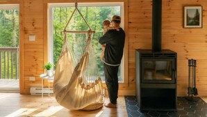 L’intérieur d’un chalet en bois, avec un hamac suspendu, un poêle à bois et une grande fenêtre donnant sur la verdure.