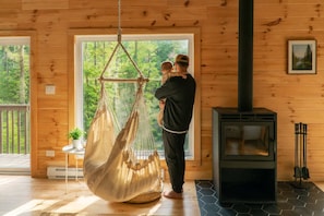 A wooden cabin interior with a hanging hammock, a wood-burning stove, and a large window overlooking greenery.