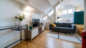 A modern loft-style room with a wooden floor, a grey sofa, a TV, a plant, and a red object. - Mercadores Apartment by Amber Star Rent (Porto)