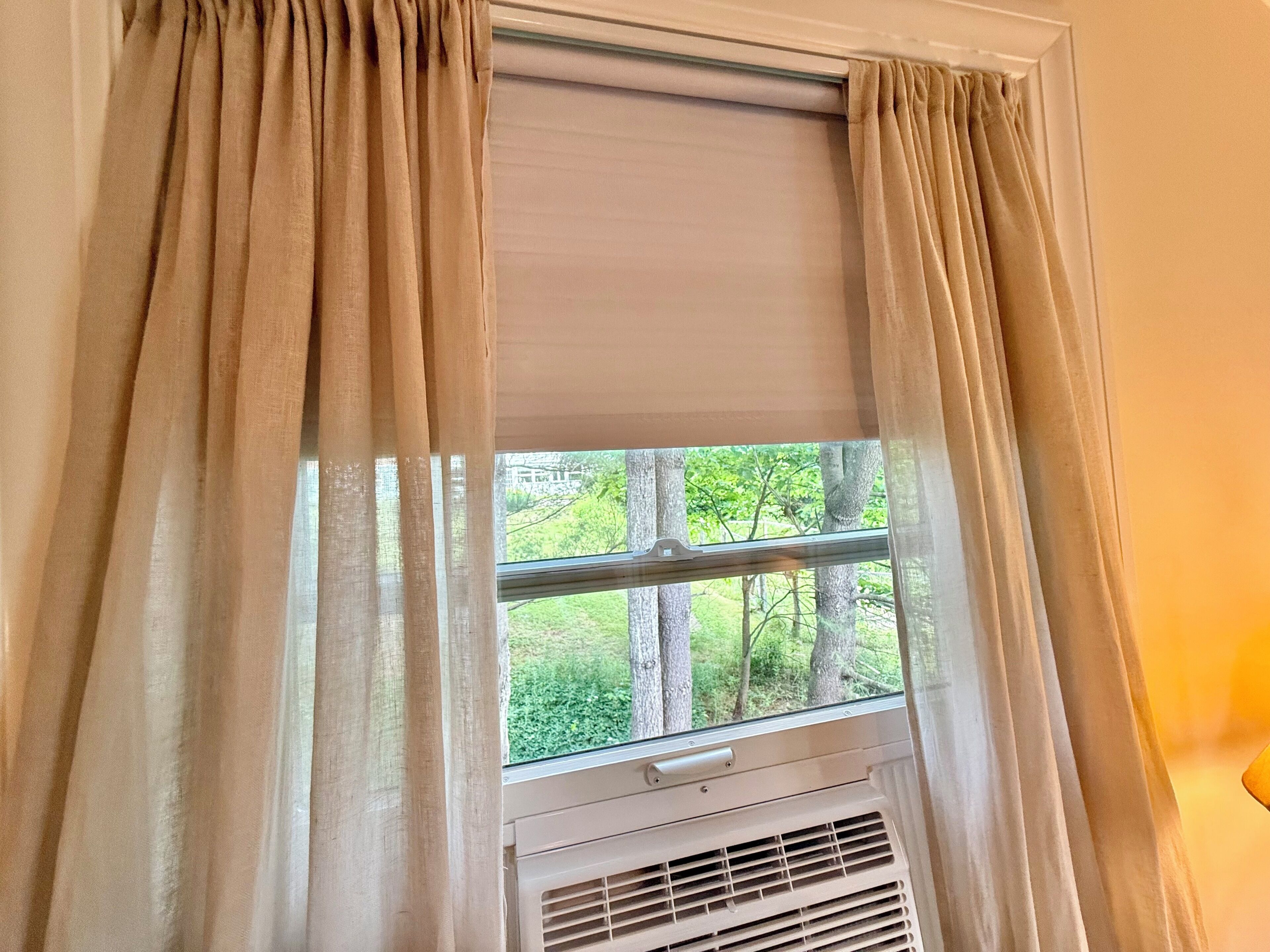 Ein Fenster mit beigen Vorhängen, Blick ins Grüne und eine weiße Klimaanlage darunter.