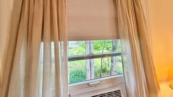 Ein Fenster mit beigen Vorhängen, Blick ins Grüne und eine weiße Klimaanlage darunter.