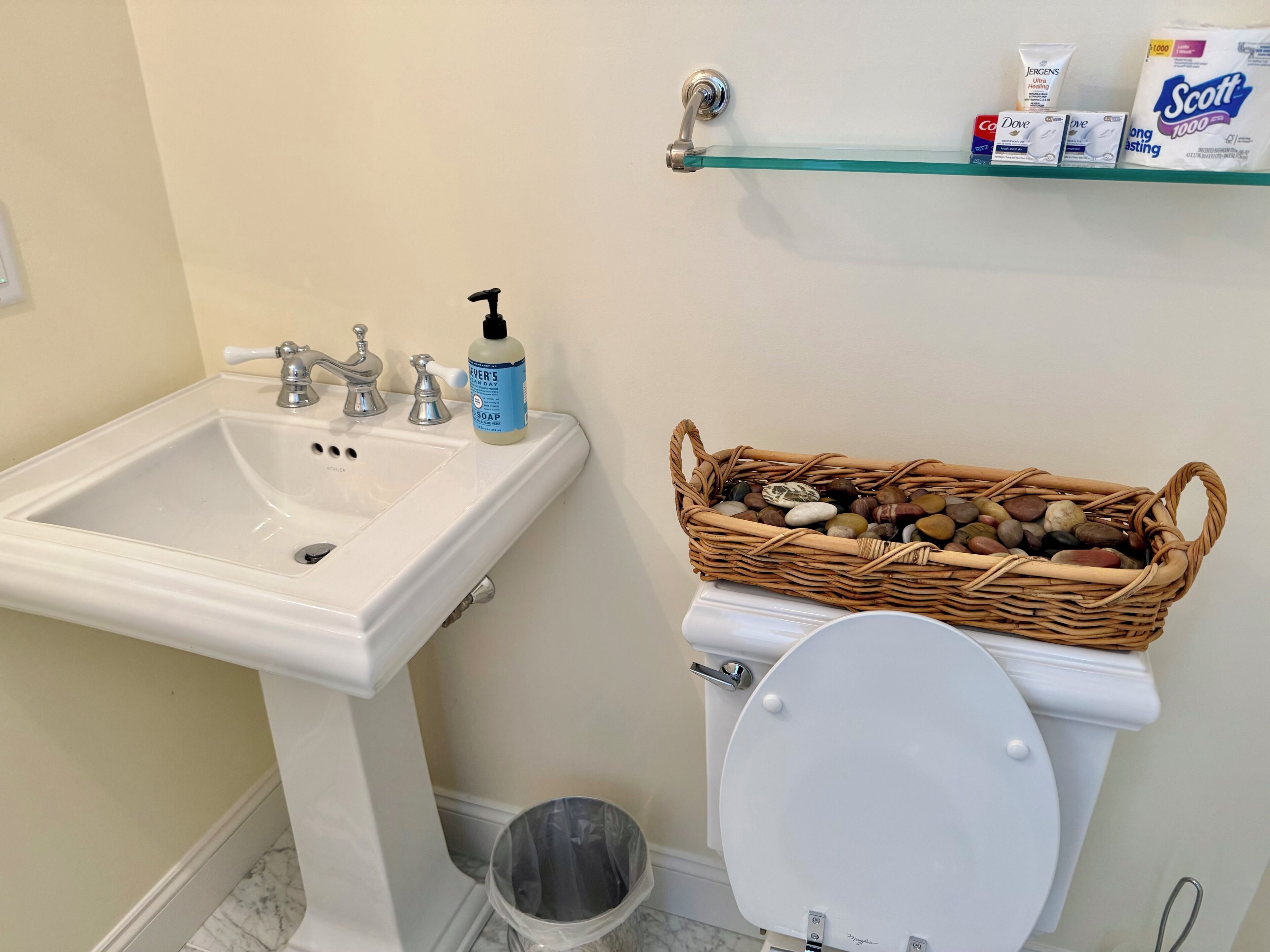 Un baño con un lavamanos blanco, un inodoro, una cesta de mimbre con piedras y artículos de aseo en un estante.