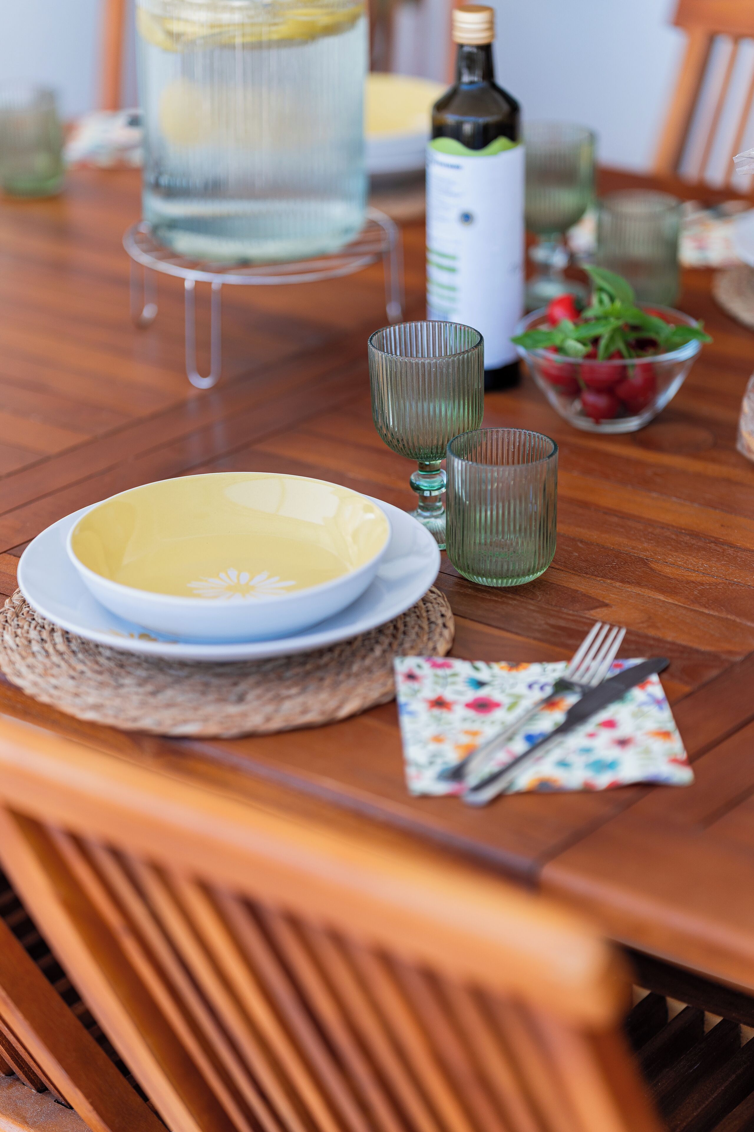 A wooden dining table with a yellow bowl, green glasses, a bottle, and a bowl of fruit.