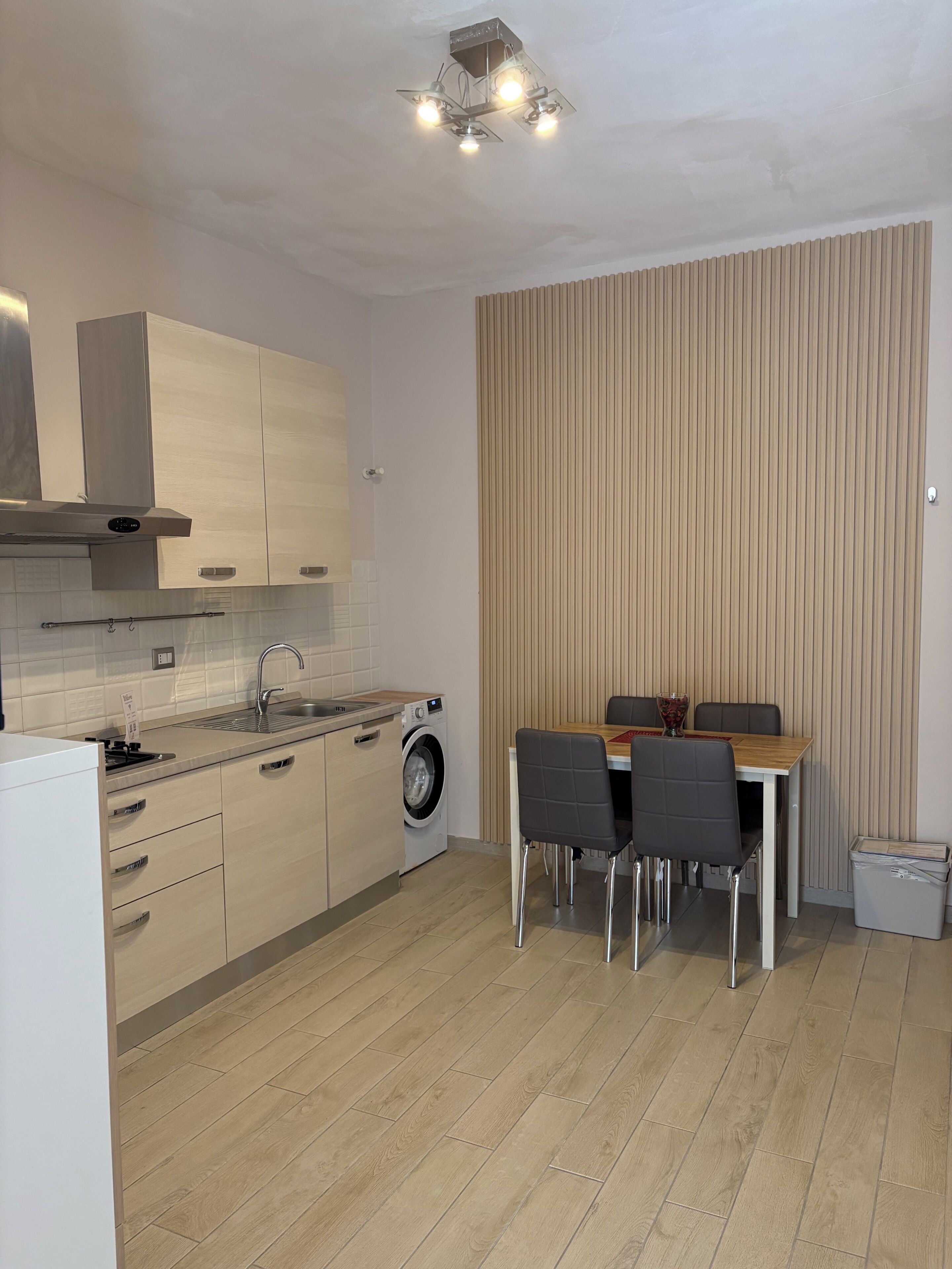 Een moderne keuken met een ingebouwde wasmachine, een eethoek met tafel en stoelen, en een wand met verticale zonwering.