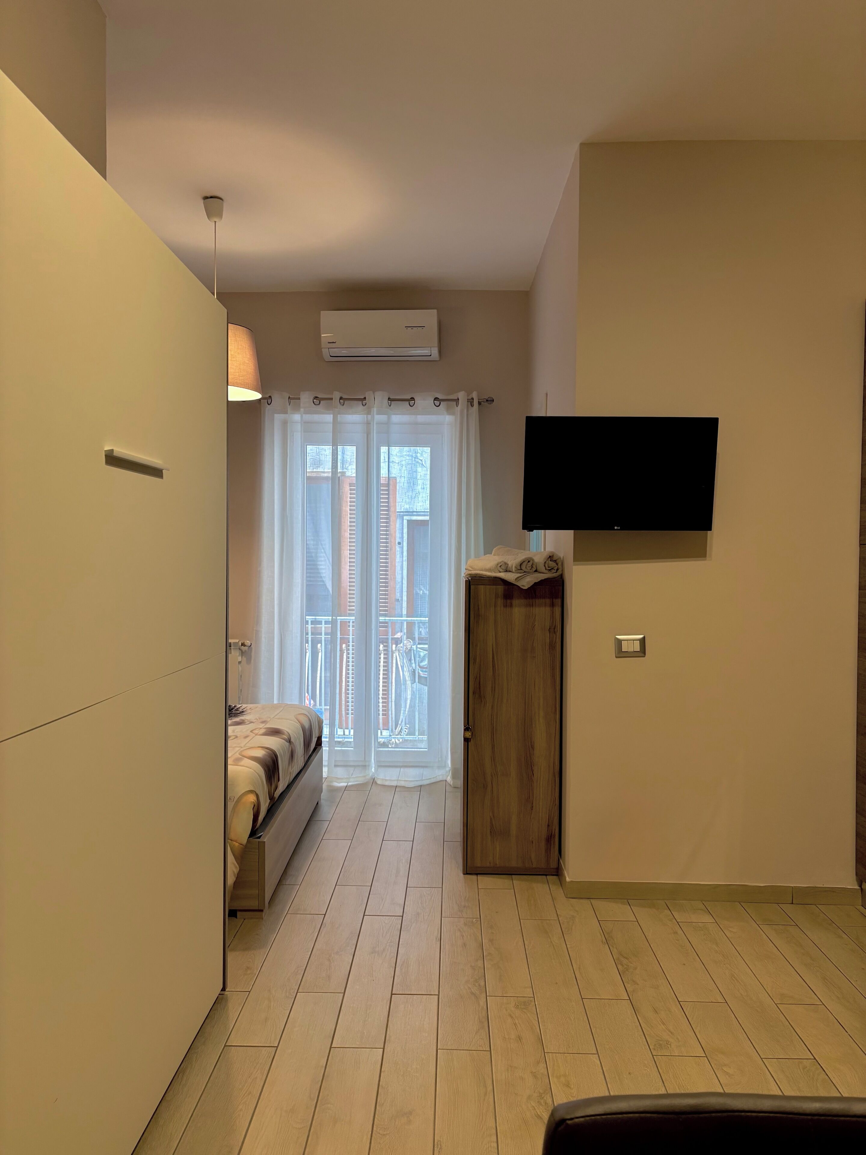 Un dormitorio con cama, televisor montado en la pared, armario de madera y puerta corrediza de vidrio hacia un balcón.
