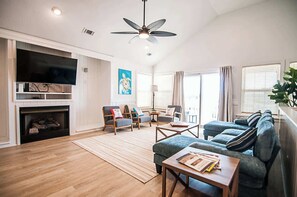 A living room with a fireplace, a television, a ceiling fan, and a large window with curtains. - Haven House-Heated Pool, Spa, Views, 1 blk to sea (Kill Devil Hills)