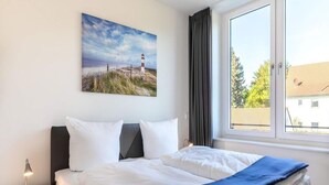 Ein Hotelzimmer mit Bett, Nachttisch, Fenster mit Vorhängen und einem Gemälde eines Leuchtturms.