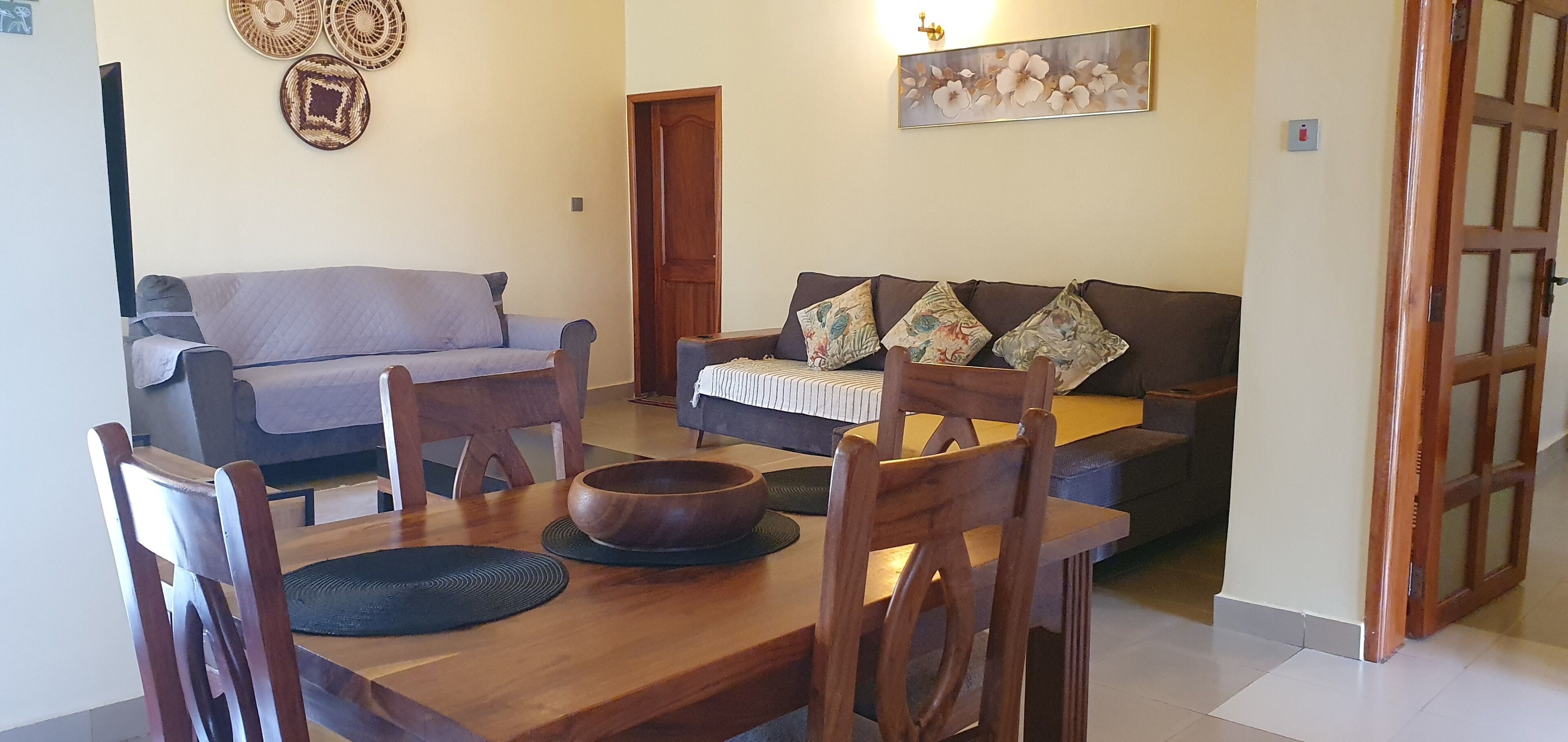 A dining area with a wooden table, chairs, and a bowl. There is a sofa and a door in the background.