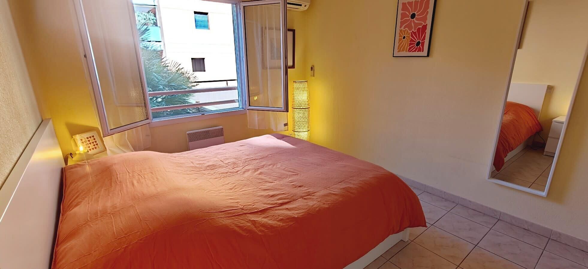 Ein Schlafzimmer mit Bett, Fenster, Heizkörper, Spiegel und einer Wand mit einem gerahmten Bild.