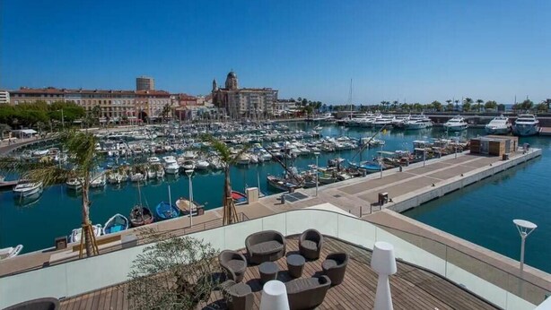 En lystbådehavn med både, en restaurant ved vandet med udendørs siddepladser, og en historisk bygning i baggrunden.