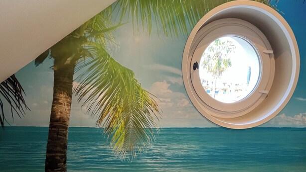 A room with a circular window overlooking a beach scene, including palm trees and the ocean. - Chic apartment between the sea and palm trees (Hyères)