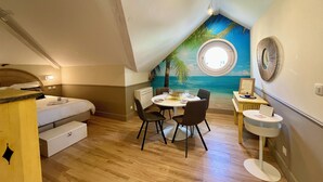 A room with a bed, a dining table with chairs, a small round table with a chair, a wall-mounted air conditioner, and a mural of a beach scene. - Chic apartment between the sea and palm trees (Hyères)