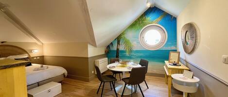 A room with a bed, a dining table with chairs, a small round table with a chair, a wall-mounted air conditioner, and a mural of a beach scene.