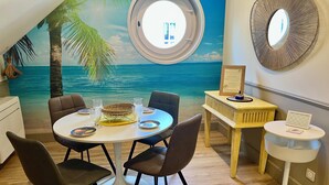 A dining area with a round table, chairs, a dessert, and a view of the ocean through a window. - Chic apartment between the sea and palm trees (Hyères)