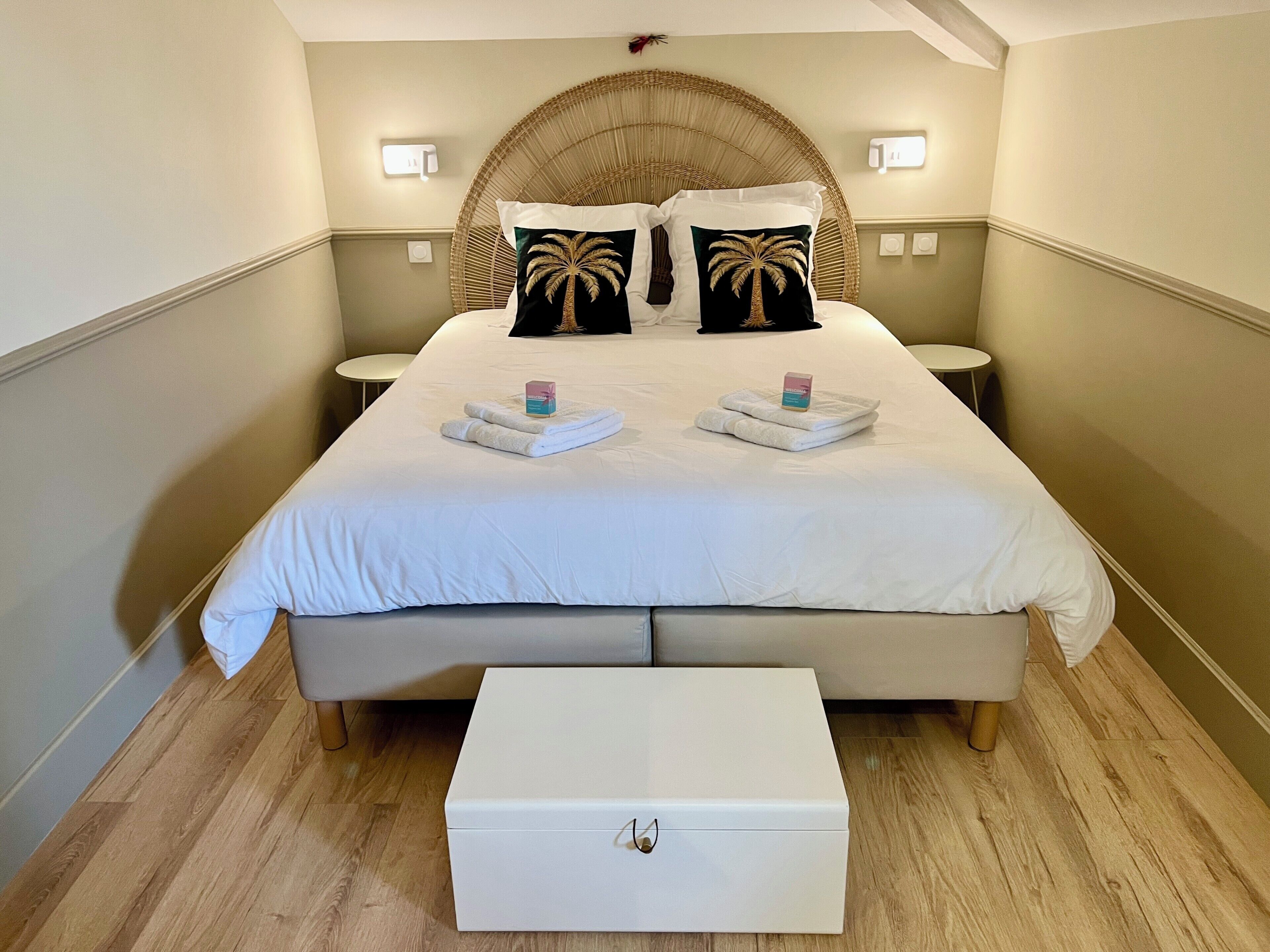 Una cama bien hecha con dos almohadas, una mesita de noche y un baúl.