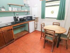 A compact kitchen with a sink, stove, microwave, and a small refrigerator. There is a dining table with chairs and a window with curtains. - Cabin No. 11 - Salt and Fern Cabins (Port Talbot)