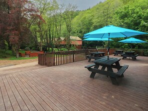 Una terraza de madera con mesas de picnic y sombrillas azules, rodeada de árboles y una valla de madera.