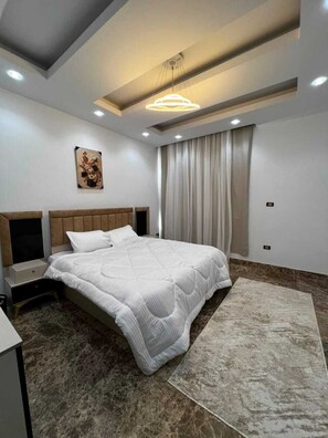 A modern bedroom with a large bed, a nightstand, a wall-mounted artwork, and a ceiling light fixture. - ELITE New Modern 3BR Apartment (New Cairo)