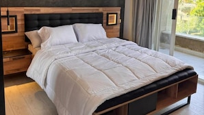 A modern bedroom with a large bed, wooden headboard, bedside lamps, and a view of greenery outside. - ELITE New Modern 3BR Apartment (New Cairo)