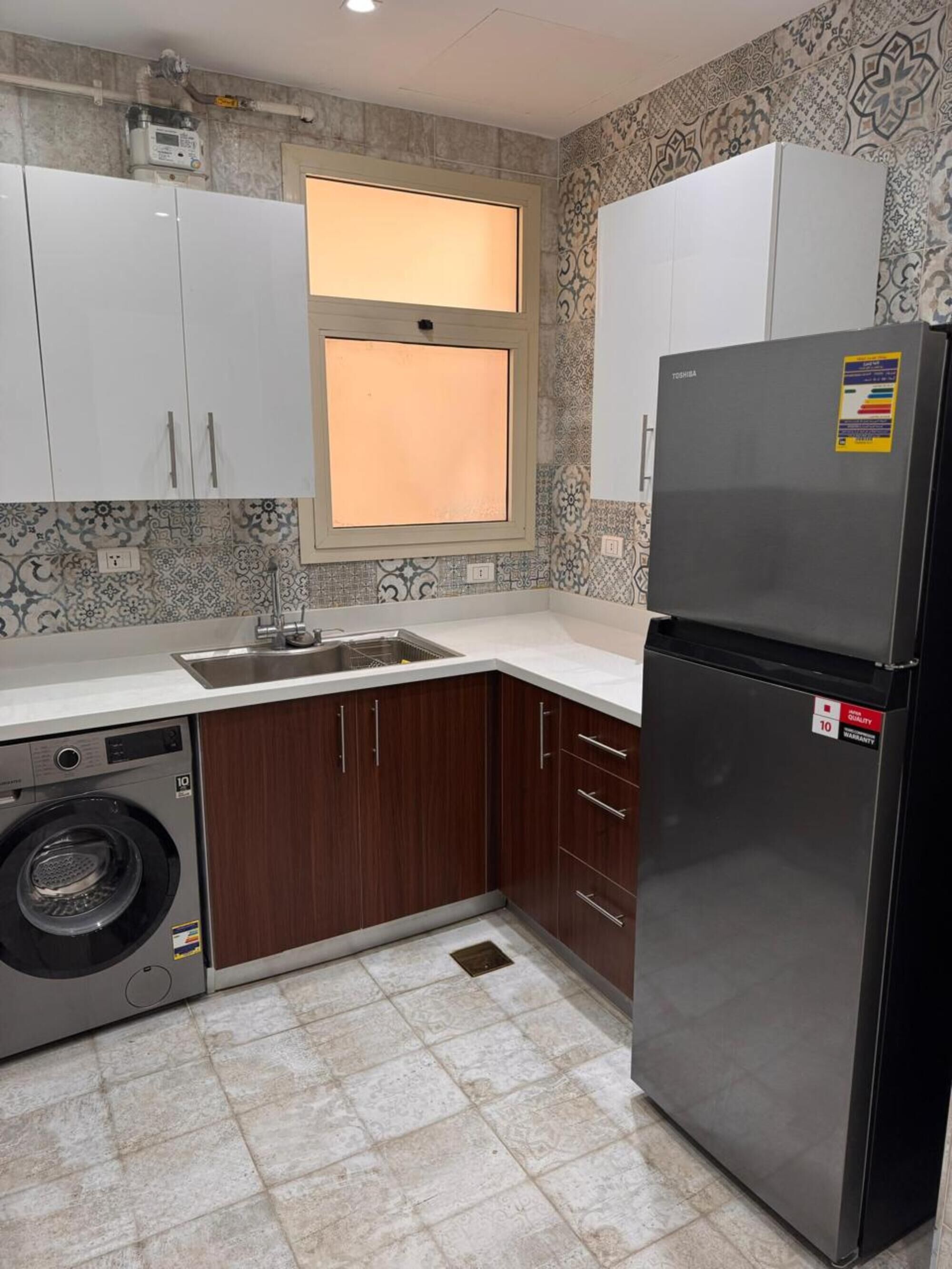 Een keuken met een wasmachine, een koelkast en een raam met verlichting.