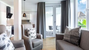 A modern living room with a sofa, armchair, and a coffee table. There is a kitchen area with a microwave and cabinets, and a balcony with a view. - 4 Star Holiday Home in Travemunde (Lübeck)