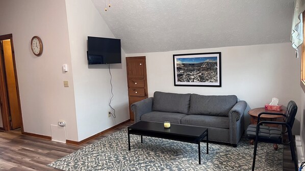 Una sala de estar con un sofá, una mesa de centro, un televisor montado en la pared, un reloj y un cuadro enmarcado.