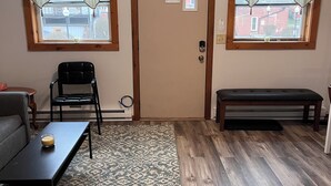 Una habitación con techo inclinado, una puerta con tres ventanas pequeñas, un banco, una mesa con un vaso y una alfombra estampada.