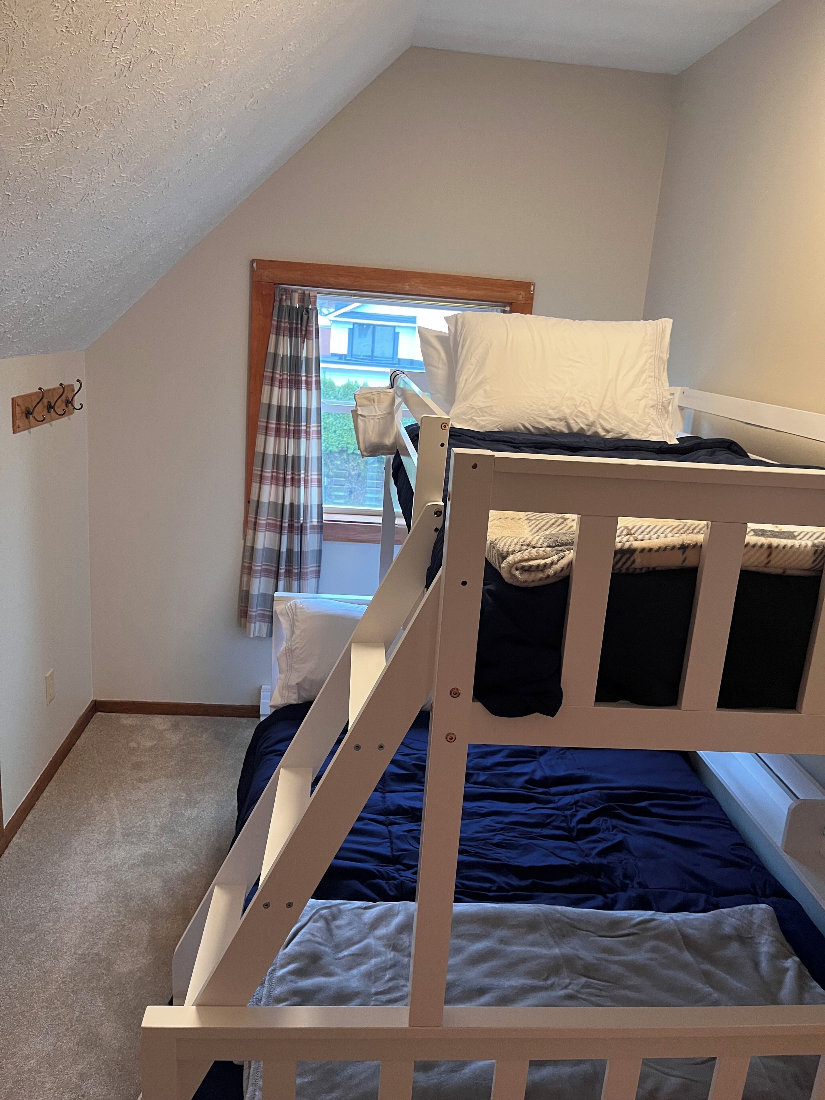 Une chambre de type dortoir, avec un lit superposé, une fenêtre avec des rideaux et une petite étagère en bois.