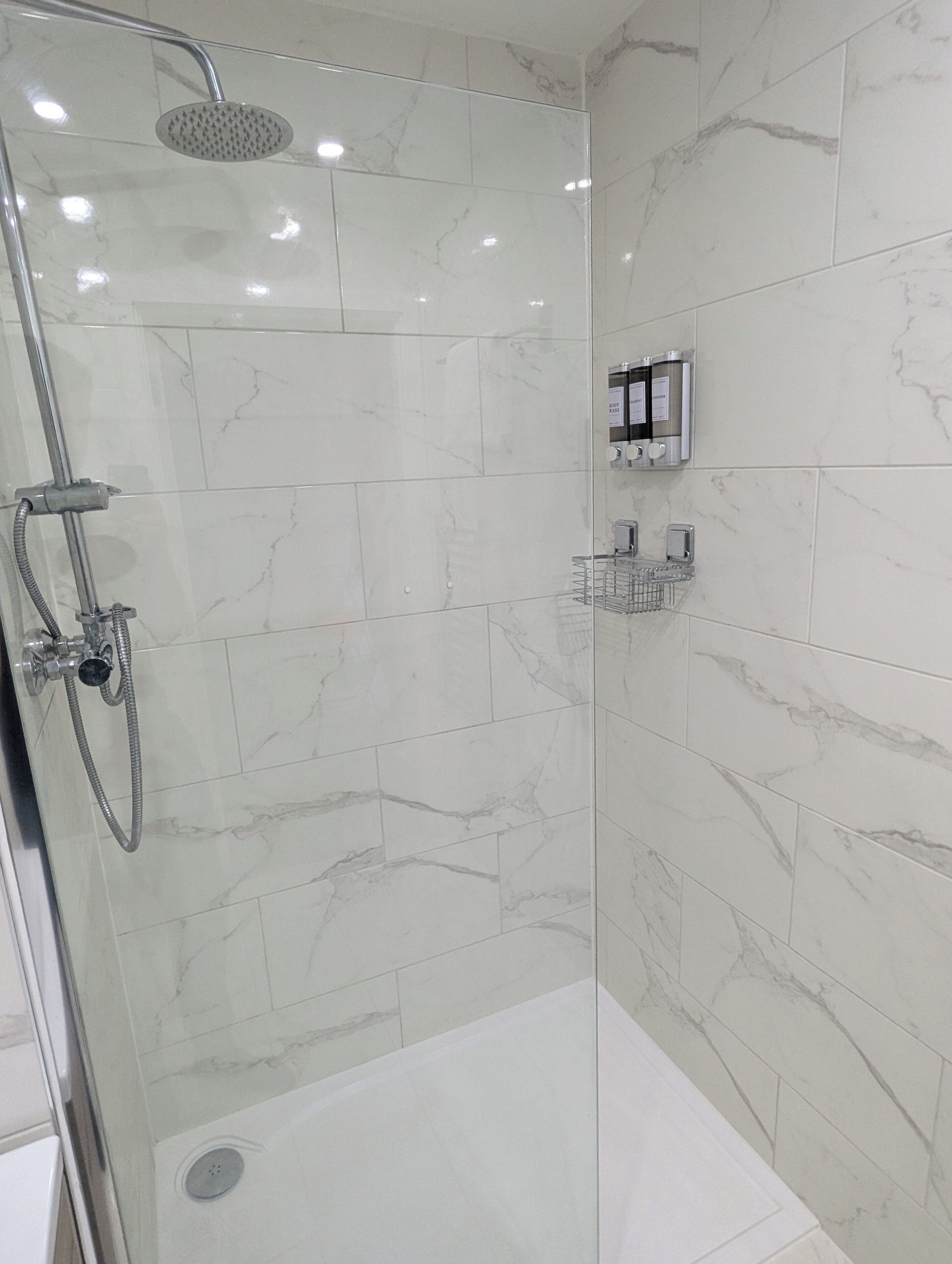 Une douche fermée par des parois en verre, dotée d’un pommeau de douche à main, d’un panneau de commande fixé au mur et d’une étagère en métal pour les articles de toilette.