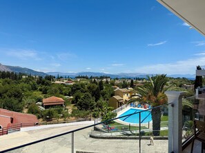 A pool with a view of mountains, a balcony with a chair, and a red-roofed building. - Thomas apartments Corfu (Corfu)