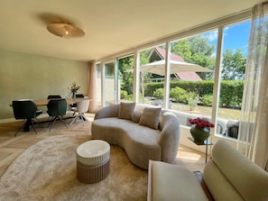 A modern living room with a curved sofa, a round ottoman, and a dining area with chairs and a table. Large windows offer a view of a house and greenery outside. - Cozy 6-guest Retreat With Fireplace (Sint Maarten)