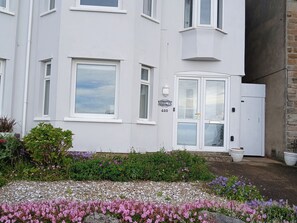 A white building with a number '480' sign, a door with glass panels, and a small garden with various flowers. - Marilyn (Morecambe)