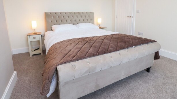 A bedroom with a tufted headboard, a brown quilted bedspread, two bedside tables with lamps, and a white door.
