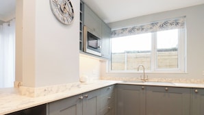 A kitchen with grey cabinets, a microwave, a clock, and a window with blinds.