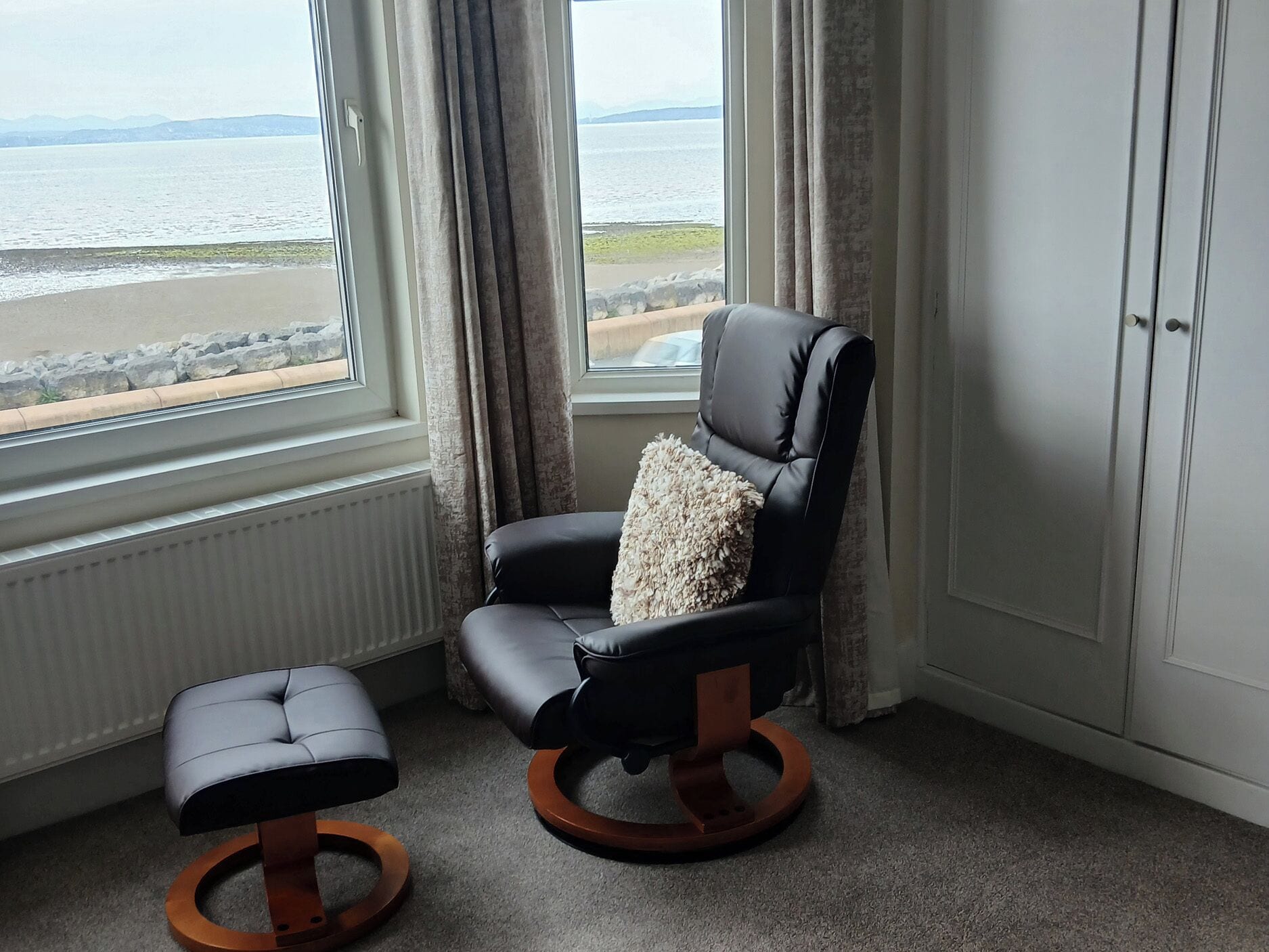 A room with a black leather chair with a cushion, a matching ottoman, and a view of the sea through a window.